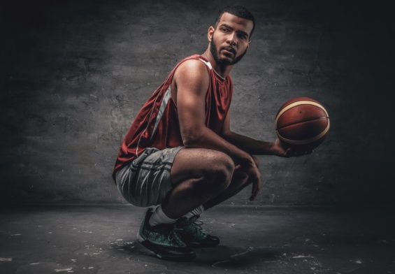 A black basketball player sits on a floor and holds basket ball.