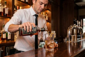 Barman versant du whisky Johnnie Walker Black Label dans un verre highball avec des glaçons sur un comptoir de bar en acajou poli